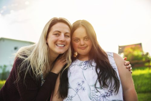 two young women one with blonde hair and dark long sleeved jumper and one with brown hair in a white singlet top with a light bue pattern smiling at camera