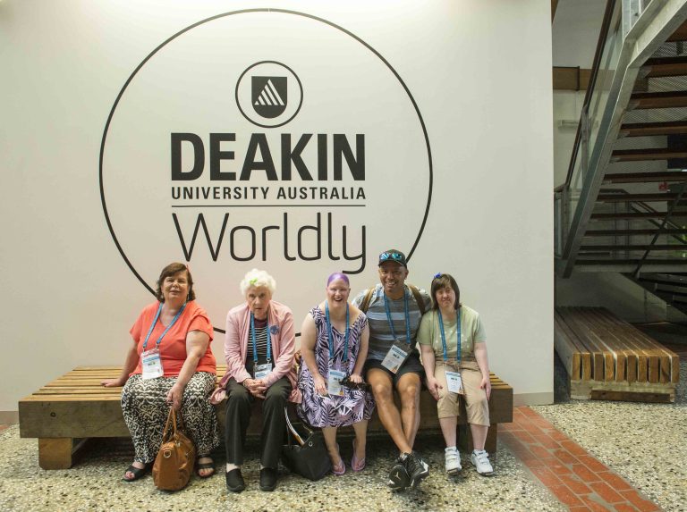 five HaS conference delegates sitting under the Deakin Uni large logo sign