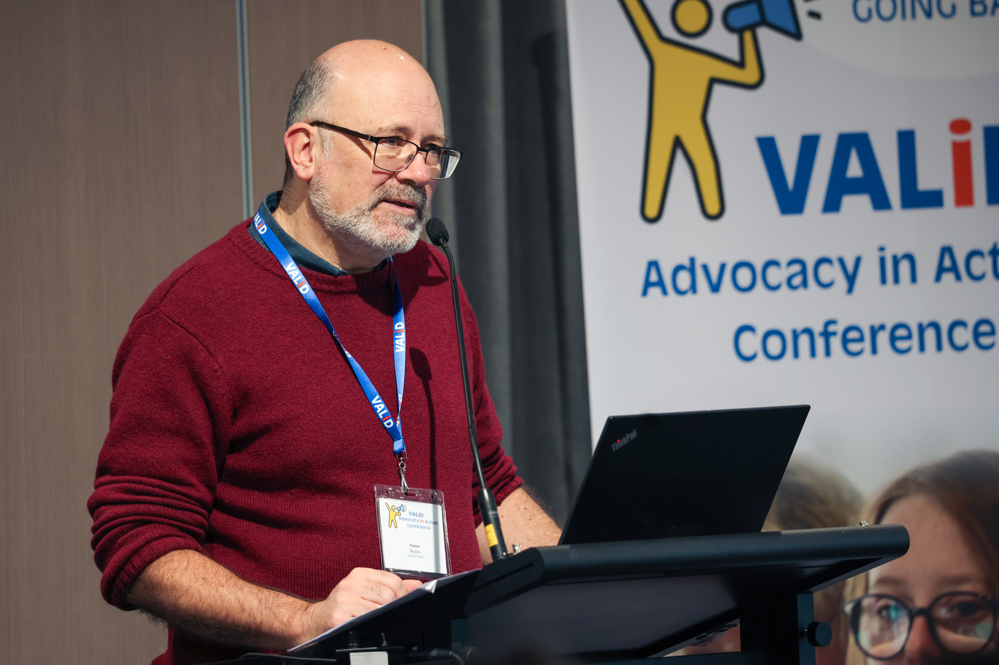 VALID CEO Fionn Skiotis, a man with fair skin, a greying bread and glasses, in speaking at a podium. He is wearing a maroon jumper and a lanyard. Behind him is a banner for the VALID Advocacy in Action conference featuring a yellow stick person with a megaphone.